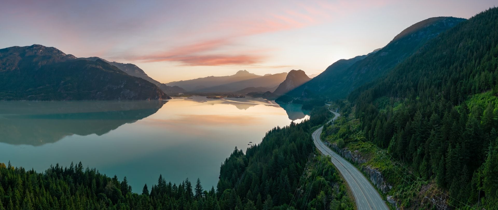 A panoramic view of a serene mountain lake reflecting a colorful sunset or sunrise sky, with a winding road alongside.