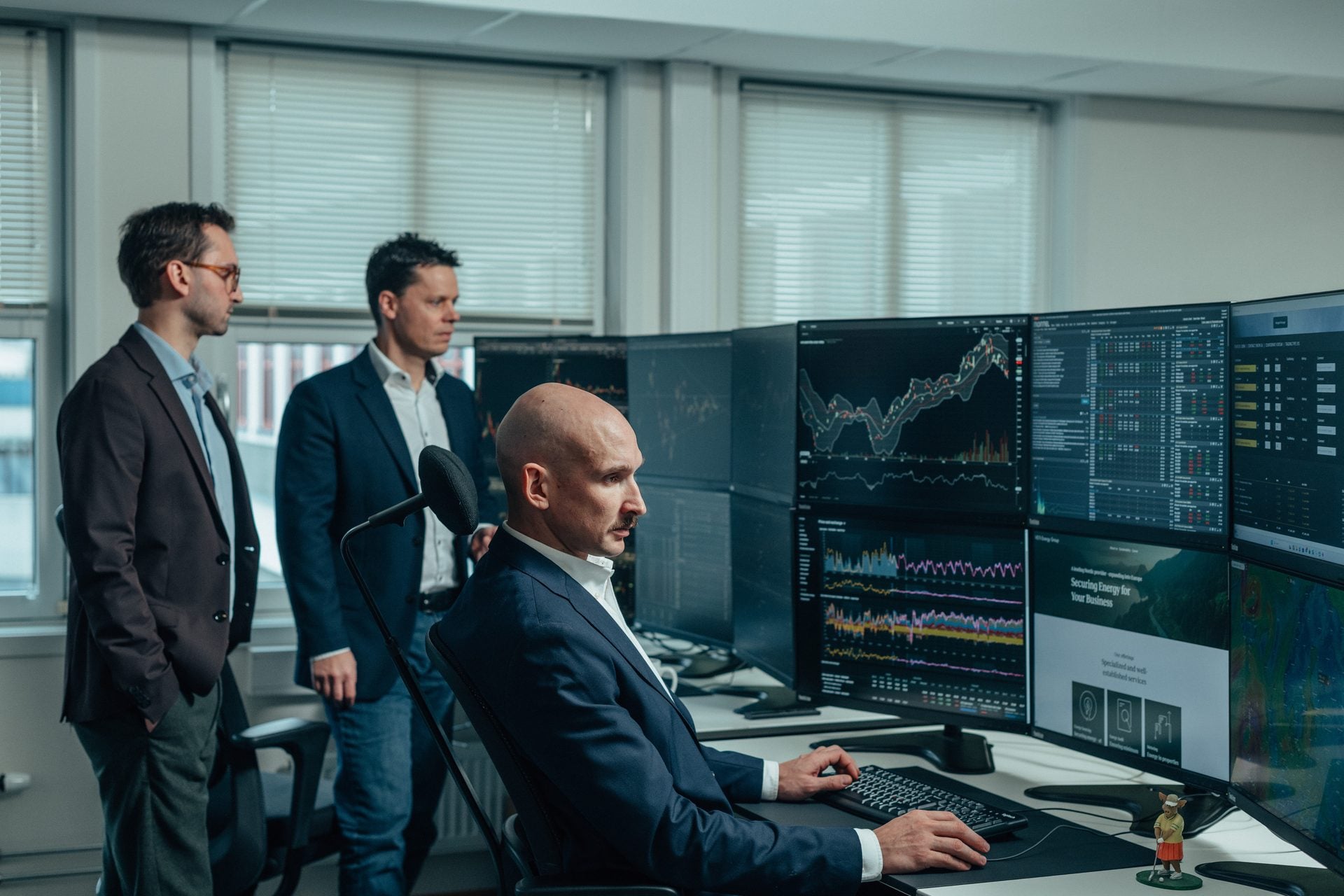 Three finance professionals, two standing and one seated, observe and analyze financial data on a multi-monitor setup in an office.